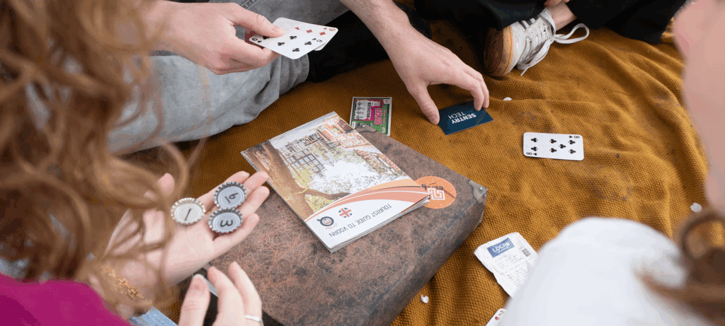 Vier vrienden zitten ontspannen op een mosterdgeel picknickkleed en spelen een gezelschapsspel: handen houden speelkaarten vast, een ander telt genummerde kroonkurken als fiches, terwijl een toeristengids, een kleine koffer en losse kaartjes tussen hen op de grond liggen.