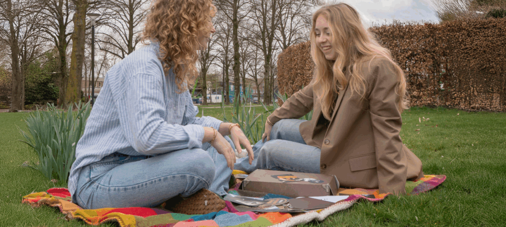 Twee jonge vrouwen zitten op een kleurrijke deken in het gras, lachend en samen een escape room spel spelend in een park, omringd door bomen in het voorjaar.